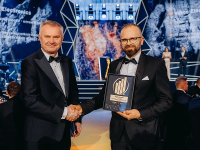 Inquadratura a media distanza di due uomini in abito da sera che si stringono la mano. L'uomo a destra, Maciej Góralski, tiene in mano un premio incorniciato con un emblema dorato che lo identifica come il premio 