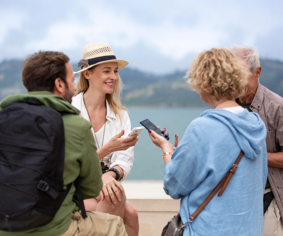 Quattro adulti, tre visibili e uno parzialmente oscurato, conversano all'aperto in riva al mare. Due donne, una delle quali sorridente e con un cappello, mostrano qualcosa sui loro traduttori che tengono in mano al gruppo.