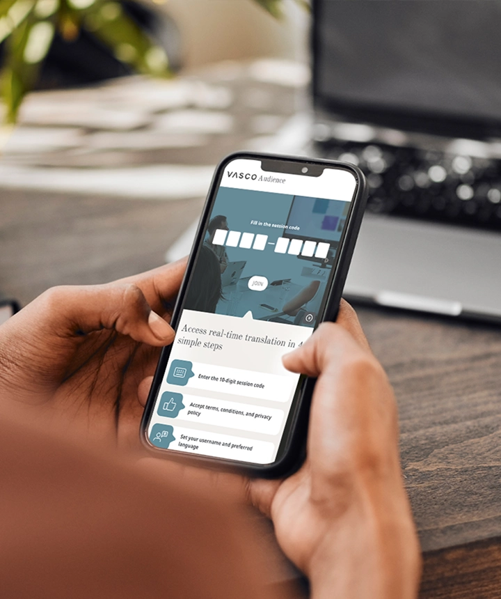 Una ripresa da dietro le spalle di una persona che tiene in mano uno smartphone su cui è visualizzata l'interfaccia web di Vasco Audience. Lo schermo mostra un campo di inserimento con la dicitura «Inserisci il codice della sessione» e un pulsante bianco «PARTECIPA», sopra un elenco di istruzioni che recita «Accedi alla traduzione in tempo reale in 4 semplici passaggi». Sullo sfondo si intravedono una scrivania di legno sfocata e un computer portatile. 
