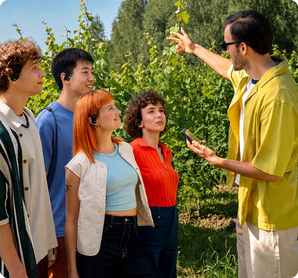 Un gruppo di cinque persone all'aperto in un vigneto illuminato dal sole. A destra, un uomo con una maglietta gialla e occhiali da sole tiene in mano un dispositivo Vasco Translator Q1 e indica con un gesto i filari di vite verdi. A sinistra, quattro persone ascoltano attentamente, ognuna con indosso un Vasco Translator E1 nero, a dimostrazione della funzione di conversazione di gruppo in tempo reale. 