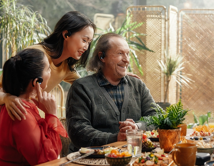 Una commovente riunione familiare multigenerazionale attorno a un tavolo da pranzo imbandito. Un uomo anziano sorridente con un cardigan grigio e due donne chiacchierano allegramente in una stanza inondata di luce e piena di piante. Tutti e tre indossano gli auricolari neri Vasco Translator E1. 