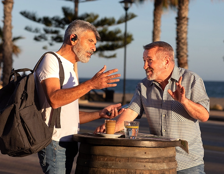 Due uomini sono in piedi accanto a un tavolo all'aperto e stanno conversando. Uno dei due ha uno zaino e entrambi indossano gli auricolari Vasco Translator E1. 