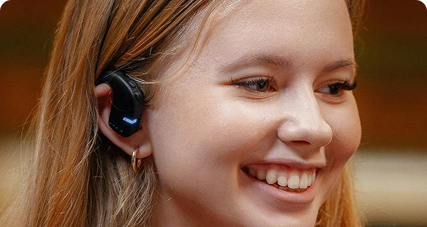 Primo piano del profilo di una giovane donna sorridente che indossa un auricolare Vasco Translation E1 nero. L'immagine mette in risalto il design igienico e aperto del dispositivo, mostrando come si adagi comodamente sulla parte esterna dell'orecchio senza entrare nel condotto uditivo. 