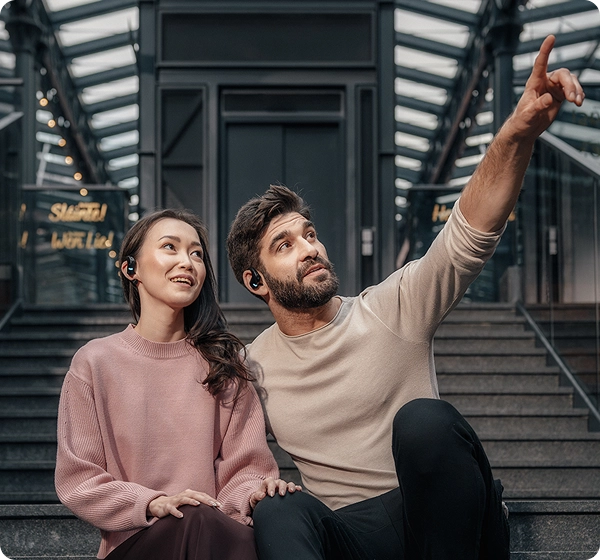 Un uomo e una donna seduti sui gradini di cemento all'interno di un edificio dall'architettura moderna con una tettoia in vetro. L'uomo indica verso l'alto con la mano, mentre entrambi indossano gli auricolari Vasco Translation E1 neri e guardano nella direzione del suo gesto. 