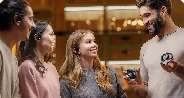 Un gruppo di quattro persone diverse che mostrano il design universale degli auricolari Vasco Translation E1 in una stanza illuminata da luci calde. Tre di loro indossano gli auricolari neri, dimostrando come si adattino comodamente a diverse forme dell'orecchio, mentre un uomo sorridente con la barba, sulla destra, tiene in mano due custodie di ricarica. 