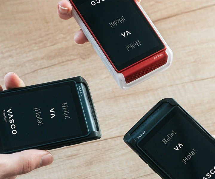 Tre persone tengono i dispositivi Vasco Translator Q1, avvicinandoli tra loro; gli schermi mostrano la schermata iniziale con il logo Vasco e le parole ‘Hello!’ e ‘¡Hola!’. I dispositivi sono presentati in custodie in silicone di tre colori: Solid Black, Dark Caramel, e Milky White.
